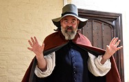 A bearded man in a red cloak, deep blue waistcoast and white shirt with billowing sleeves wears a wide-brimmed grey hat and gestures with hands open